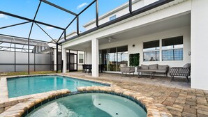 A swimming pool with a hot tub, surrounded by a covered patio area with seating and a view of the sky.