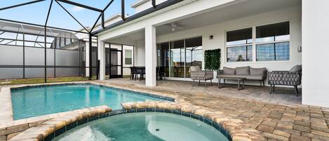 A swimming pool with a hot tub, surrounded by a covered patio area with seating and a view of the sky.