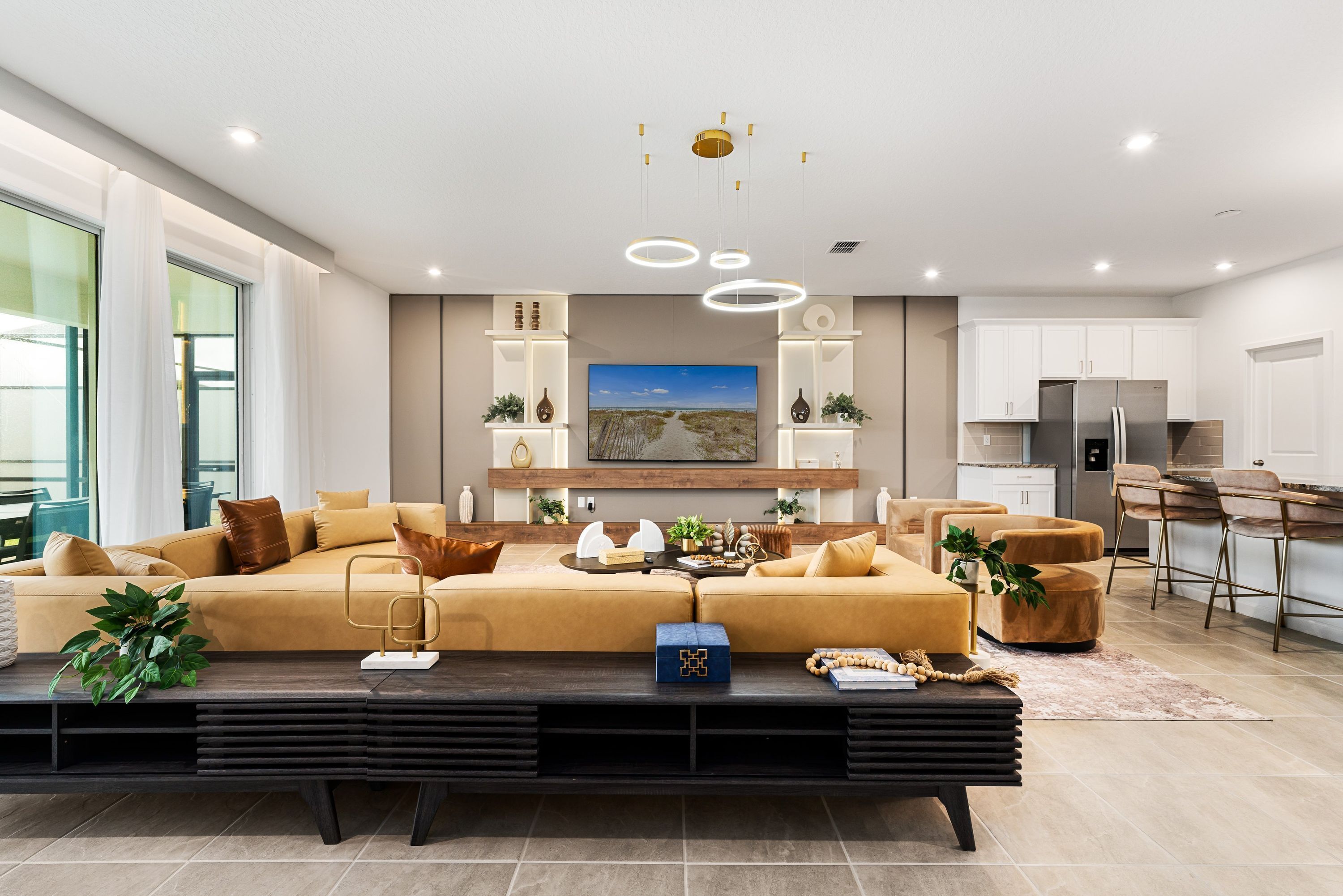 A modern living room with a large sofa, a coffee table, and a TV. There is a kitchen area with a refrigerator and cabinets.