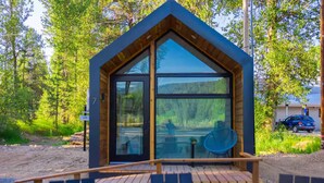 Una cabaña pequeña con un gran ventanal, una terraza de madera y vistas al bosque circundante.
