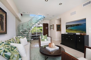 A modern living room with a staircase, a large flat-screen TV, and a view of the outdoors. - Kapalua Bay | Ocean View 4 BR, Sleeps 14 | Car Incl. w/ 6+ Nights | KBV ML-2080 by KBM (Lahaina)