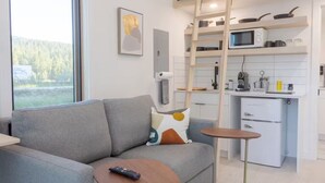 Una sala de estar moderna con un sofá gris, una mesa de centro y un control remoto encima. Hay una zona de cocina con armarios blancos, un microondas y una cafetera. Una escalera está apoyada contra la pared y hay una ventana con vistas a árboles.