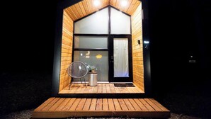 Una cabaña de madera pequeña con interior oscuro, una sola puerta y un porche con una silla y una maceta.