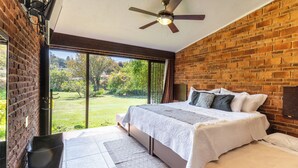Une chambre avec un grand lit, un ventilateur de plafond, un mur en briques et une vue sur un paysage verdoyant, visible à travers une porte coulissante.