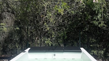 A hot tub with a wooden exterior, surrounded by dense foliage and a metal fence.