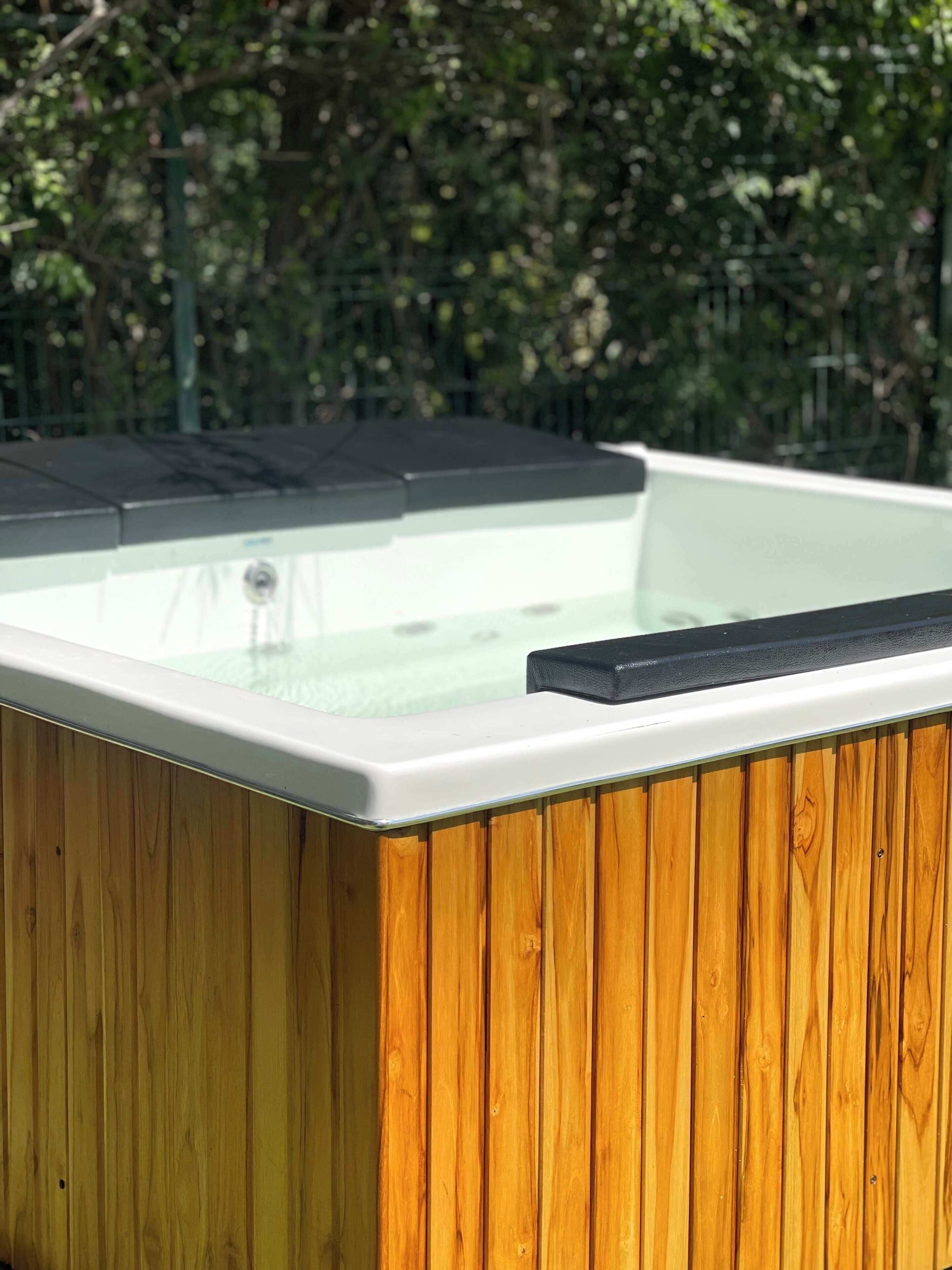 A hot tub with a wooden exterior and a glass top, surrounded by greenery.