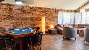 Une pièce chaleureuse avec un mur en briques, une table en bois sur laquelle est posé un jeu de société, des sièges confortables et une grande fenêtre avec des rideaux.