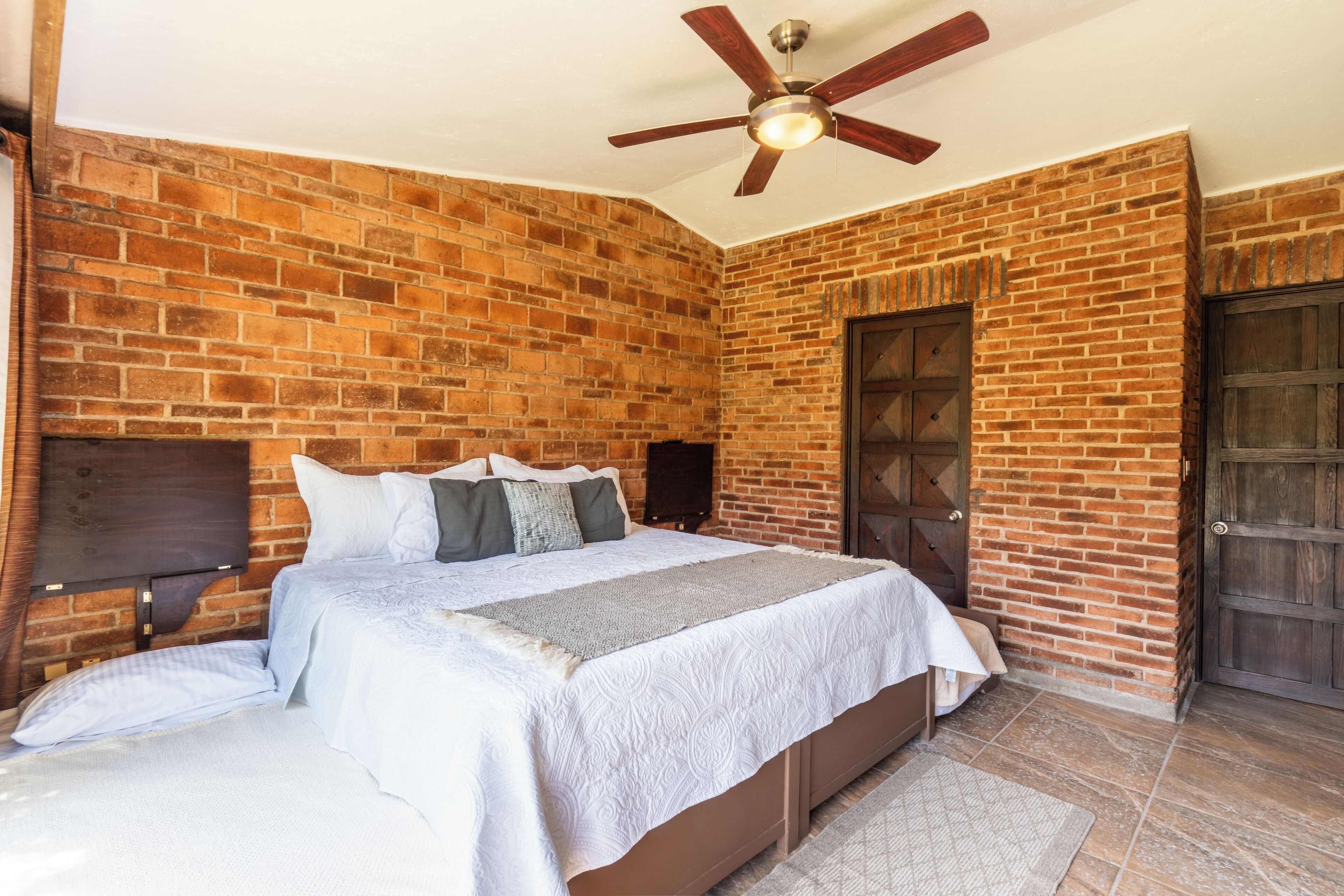 A bedroom with a brick wall, a bed with white bedding, a ceiling fan, and a wooden door.