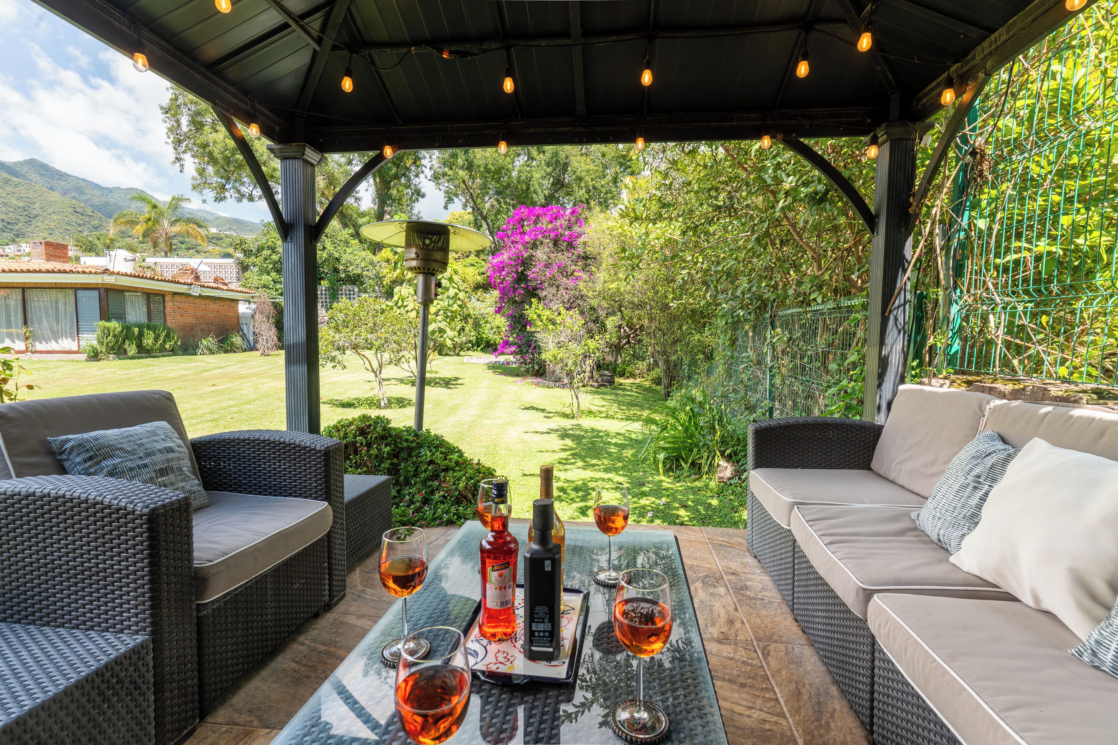 Un patio exterior con mobiliario de mimbre, una mesa con vasos y botellas, y vistas a un jardín y montañas.