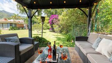 Un patio exterior con mobiliario de mimbre, una mesa con vasos y botellas, y vistas a un jardín y montañas.
