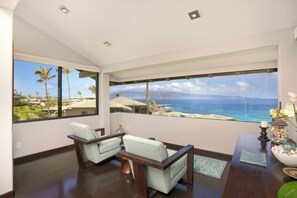 Ein Zimmer mit einem großen Fenster, das Blick auf das Meer und Palmen bietet. Zwei Sessel und ein kleiner Tisch mit Dekorationen sind vorhanden.