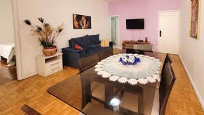 A living room with a dark blue sofa, a white coffee table with a blue centerpiece, a flat-screen TV mounted on the wall, and a wooden floor. - Copa Beach Walk – 3 Bedrooms,5 minutes from the beach • Air conditioning + Wi-Fi (Rio de Janeiro)