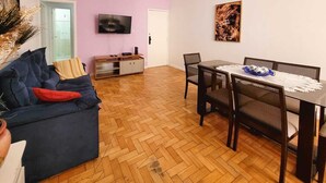 A living room with a sofa, a dining table with chairs, a TV mounted on the wall, and a door leading to another room. - Copa Beach Walk – 3 Bedrooms,5 minutes from the beach • Air conditioning + Wi-Fi (Rio de Janeiro)