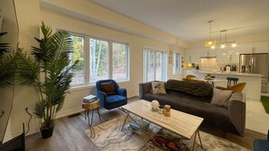 A modern living room with a sofa, coffee table, and a dining area. There are large windows providing natural light and a view of the outdoors. - Luxury Muskoka Beach Retreat, Games Room & Hot Tub (Gravenhurst)
