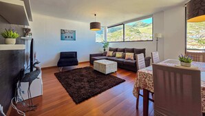 Ein modernes Wohnzimmer mit einer Couch, einem Couchtisch und einem Essbereich mit Tisch und Stühlen. Ein großes Fenster bietet Blick auf ein Dorf am Hang.