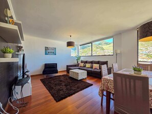Ein modernes Wohnzimmer mit einer Couch, einem Couchtisch und einem Essbereich mit Tisch und Stühlen. Ein großes Fenster bietet Blick auf ein Dorf am Hang.
