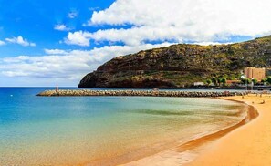 A beach with clear water, a rocky breakwater, and a building on a hillside. - Oceanfront Apt w/ Balcony & Free Parking, Machico (Machico)