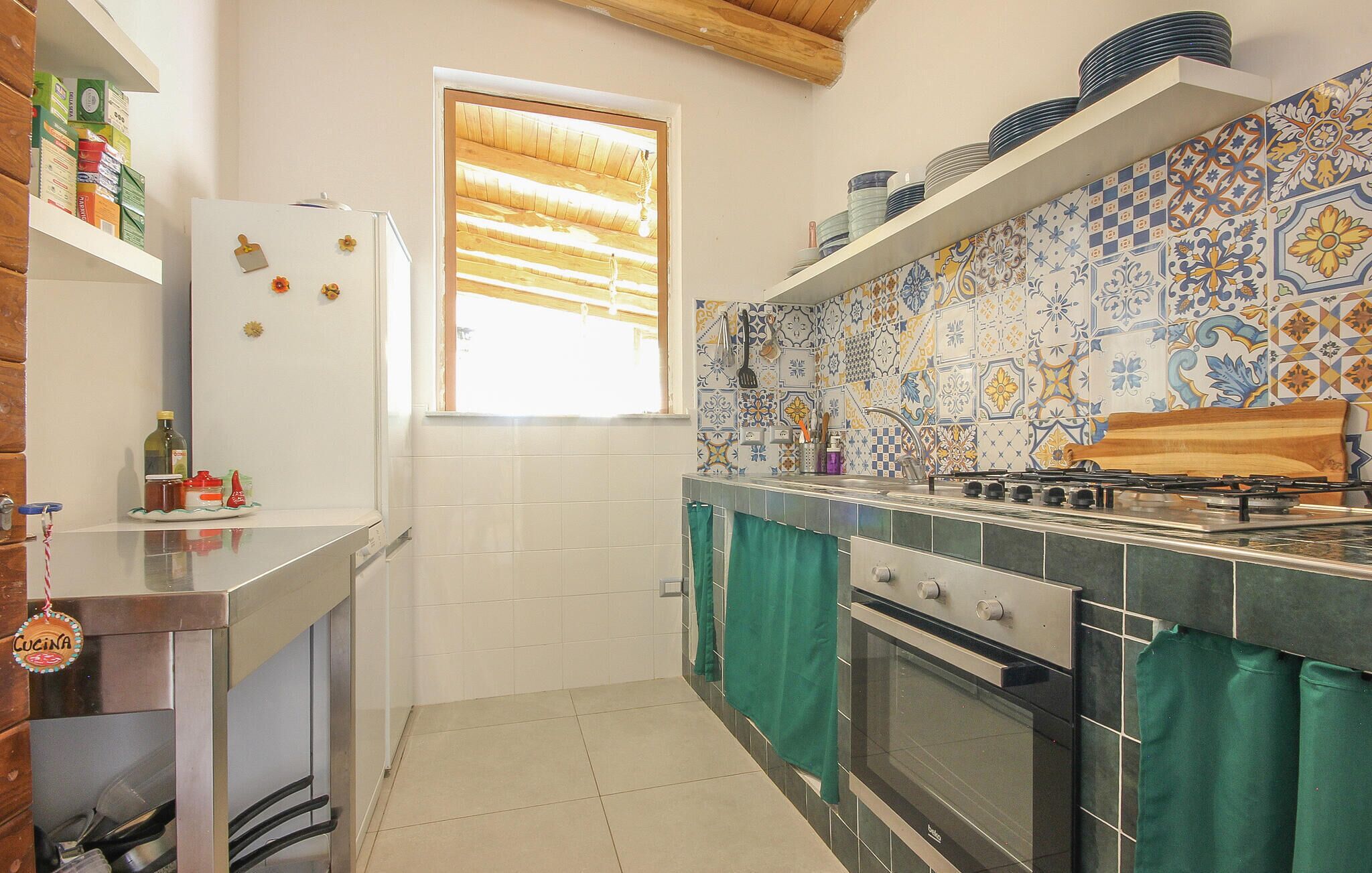 A compact kitchen with a refrigerator, oven, and stove, tiled backsplash, and a window with blinds.