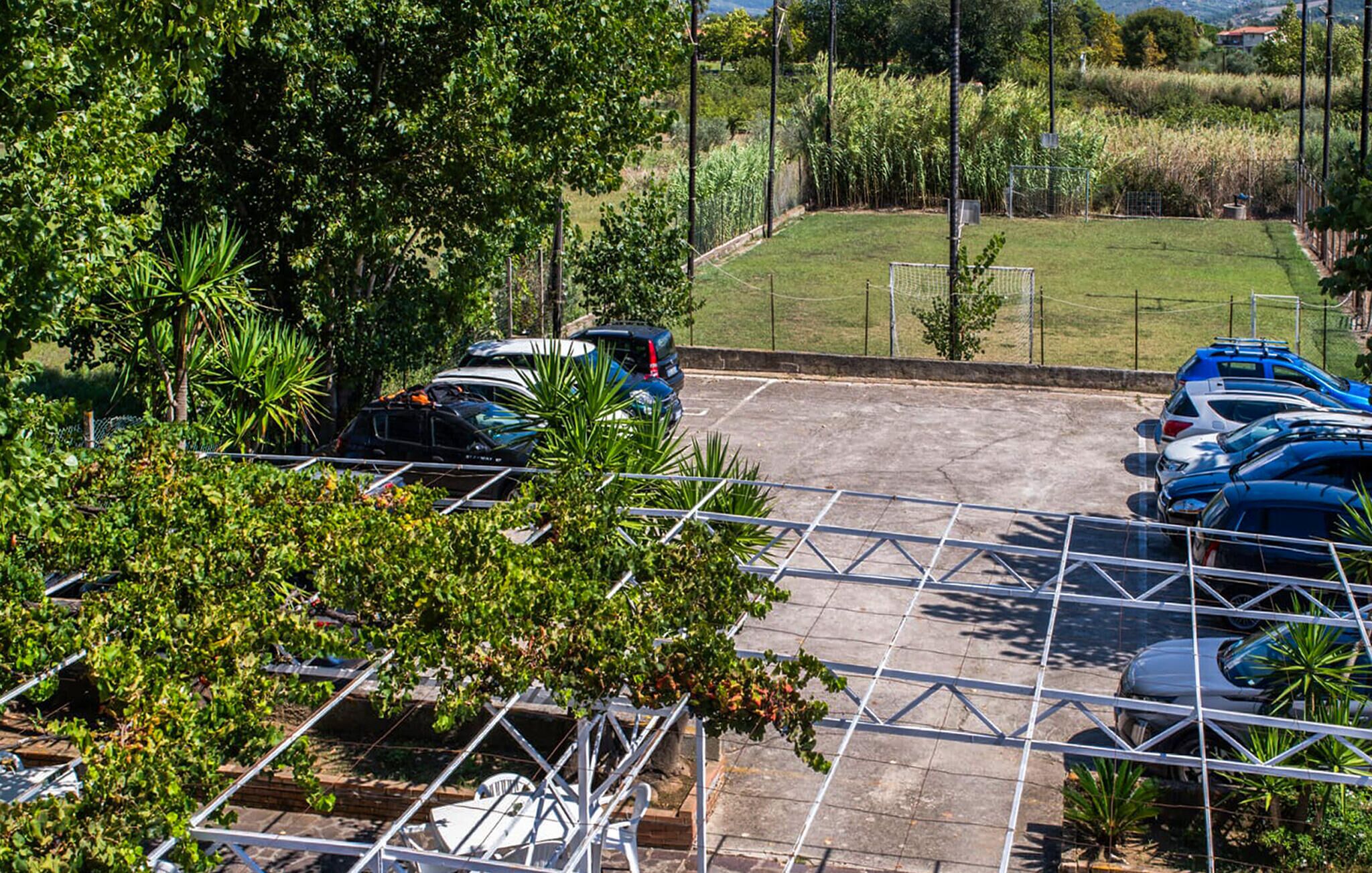 Zona d'aparcament amb diversos cotxes, envoltada de vegetació i un camp esportiu tancat.