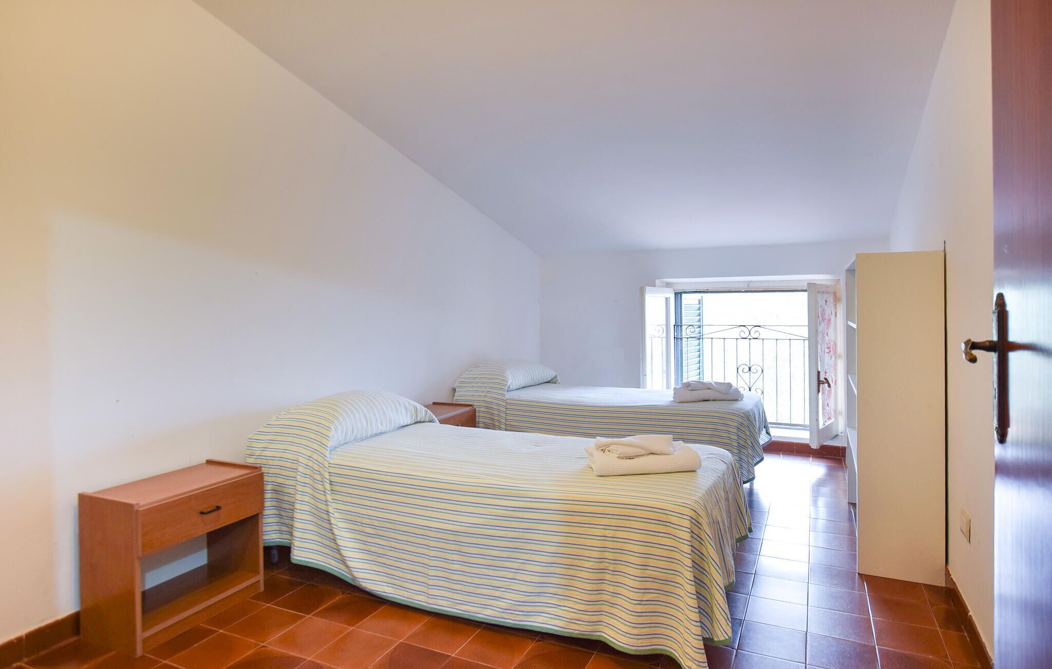 Quarto pequeno e limpo com duas camas, uma mesa de cabeceira de madeira e uma porta para uma varanda.