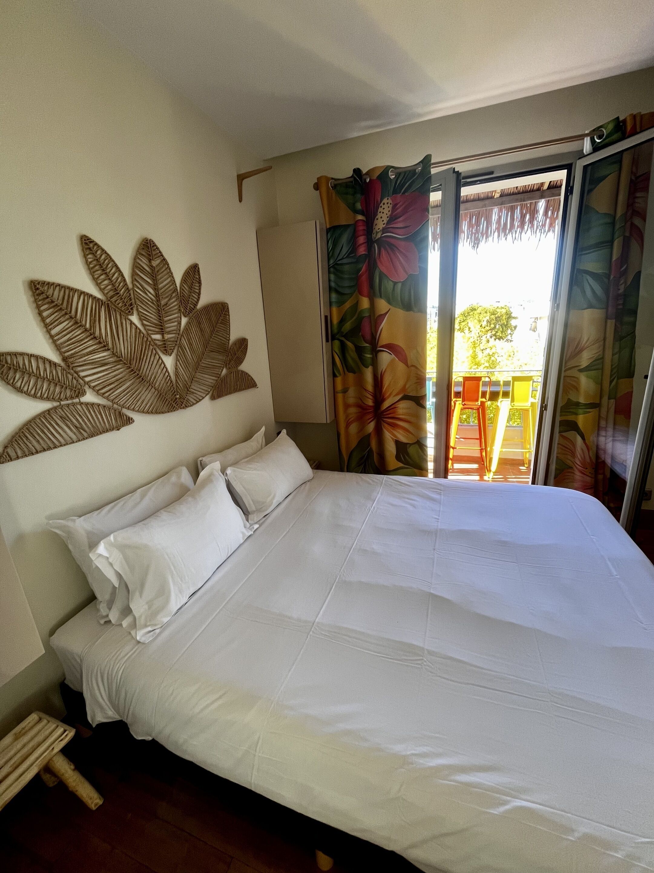 A neatly made bed with white linens in a room with a tropical leaf wall art, a sliding glass door to an outdoor area, and a wooden stool by the bed.