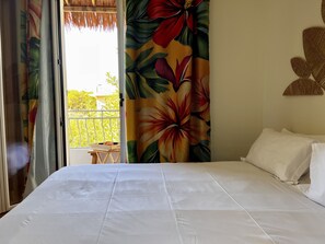 Hotellihuone, jossa on sänky, valkoiset verhot ja näkymä parvekkeelle, jossa on pöytä ja tuolit.