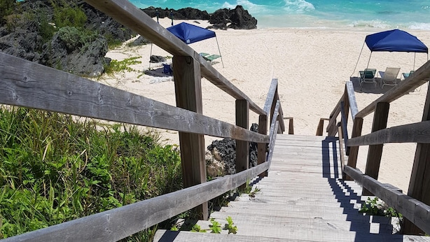 Een houten trap die naar een strand leidt, met blauwe parasols en stoelen.