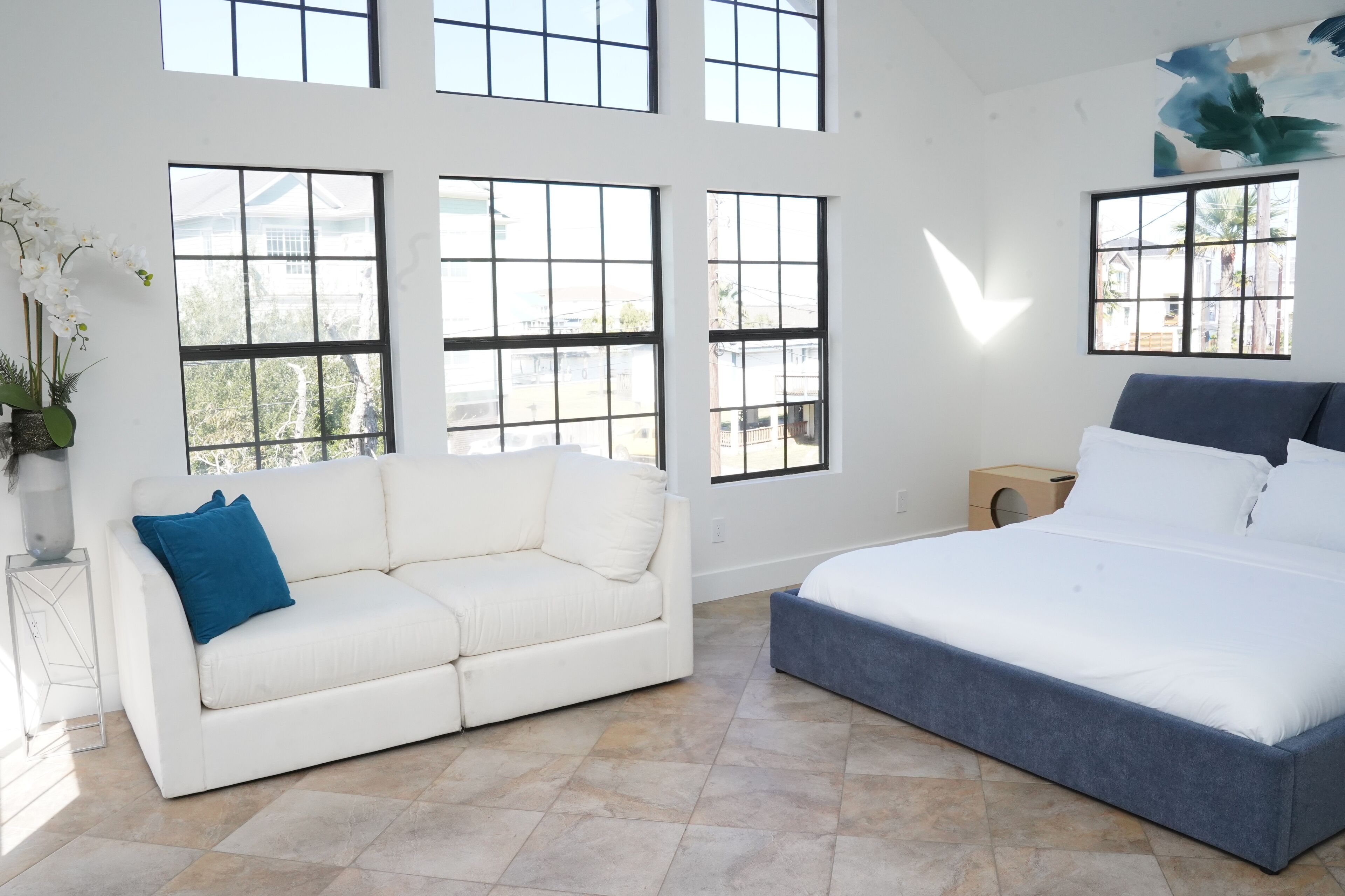 Ein modernes Schlafzimmer mit einer weißen Couch, einem großen Bett und mehreren Fenstern mit Blick ins Freie.
