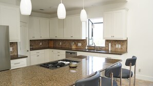 Een moderne keuken met witte kasten, granieten aanrechtbladen, een centraal eiland met barkrukken en hanglampen.