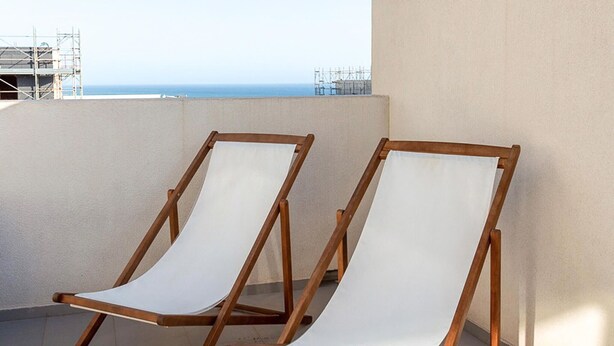 Two wooden lounge chairs with white cushions on a balcony with a view of the sea and construction in the background. - Cozy apartment in Ragusa with house sea view (Ragusa)