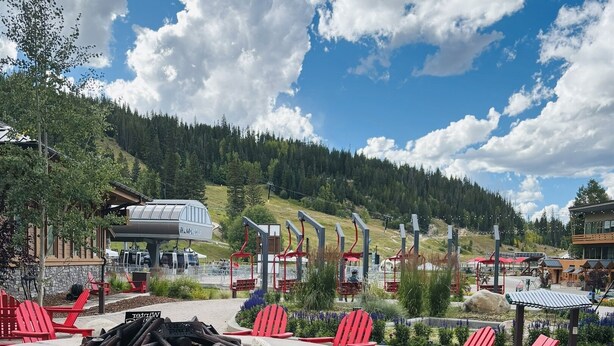 Ein runder Feuerkorb, umgeben von roten Adirondack-Stühlen, mit einer Seilbahn und einer Berghütte im Hintergrund.