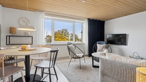 Uma sala de estar moderna com área de jantar, TV na parede e uma grande janela com vista para árvores.