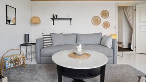 Uma sala de estar moderna com um sofá cinzento, uma mesa de centro redonda, uma mesinha lateral preta, um suporte de revista em vime e uma prateleira fixada na parede com objetos decorativos.