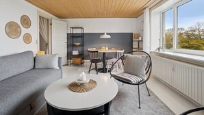 Uma sala de estar moderna com um sofá cinza, uma mesa redonda branca, uma cadeira preta e uma grande janela com vista.