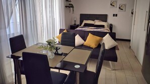 Ein modernes Hotelzimmer mit einem Bett, einem Esstisch mit Stühlen, einem kleinen Beistelltisch und einem großen Fenster mit leichtem Vorhang.