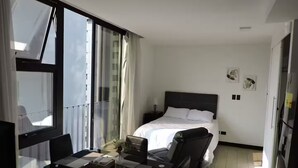 Ein modernes Hotelzimmer mit großem Fenster, einem Bett mit weißer Bettwäsche, einem Schreibtisch mit Laptop und einem Sessel.