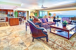 A spacious living area with a large window offering a view of the ocean, a kitchen with modern appliances, and a cozy seating area with plush sofas and a coffee table. - Honua Kai | Ocean Front 3 BR | Free Car w/ 6+ Nights | HKH-501 (Lahaina)