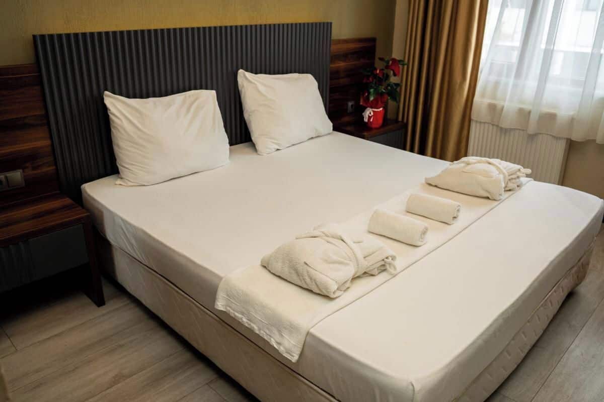 Una cama bien hecha con almohadas, una mesita de noche con un florero y una ventana con cortinas.
