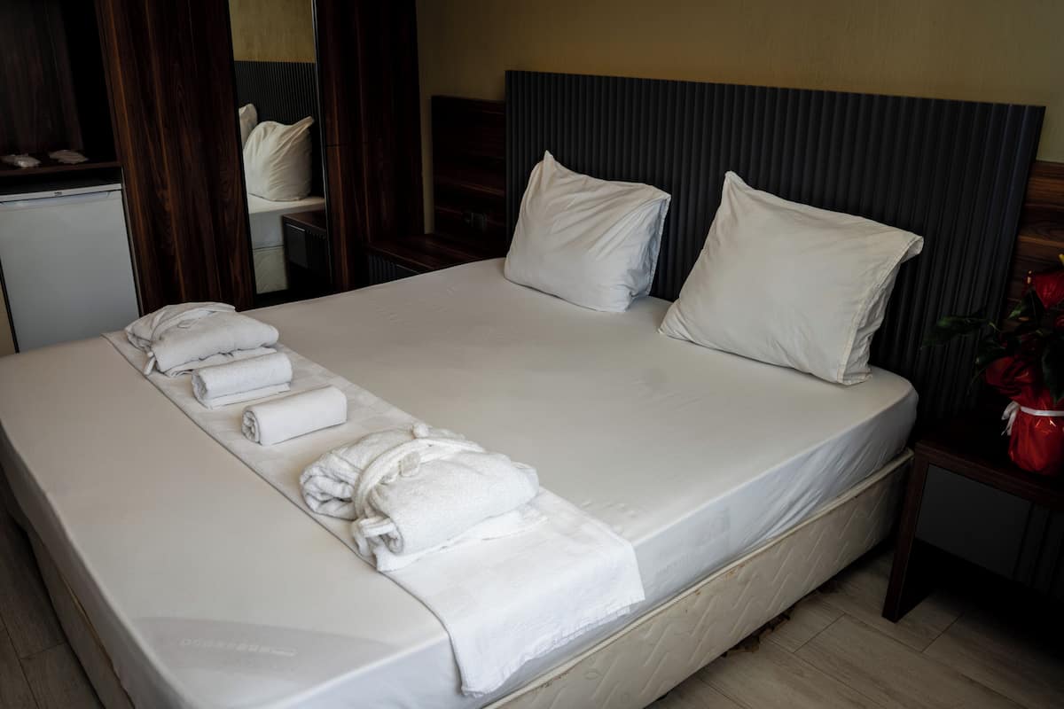 Una cama bien hecha con sábanas y toallas blancas, un cabecero de madera y una mesita de noche con un jarrón rojo.
