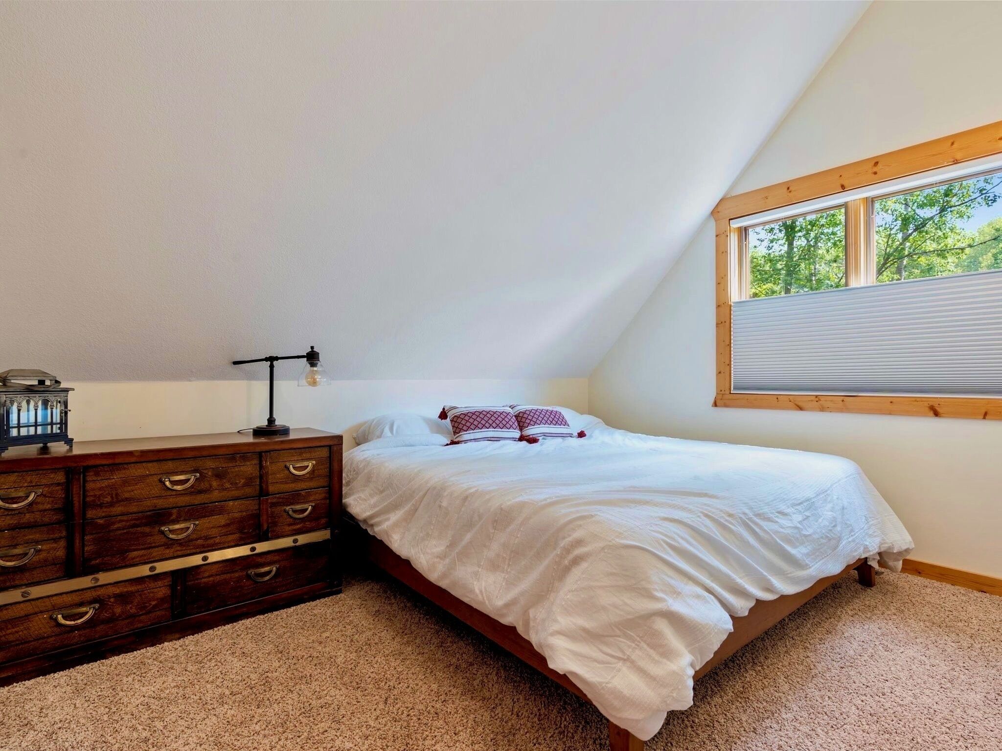 A bedroom with a bed, a dresser, a lamp, and a window with a view of trees.