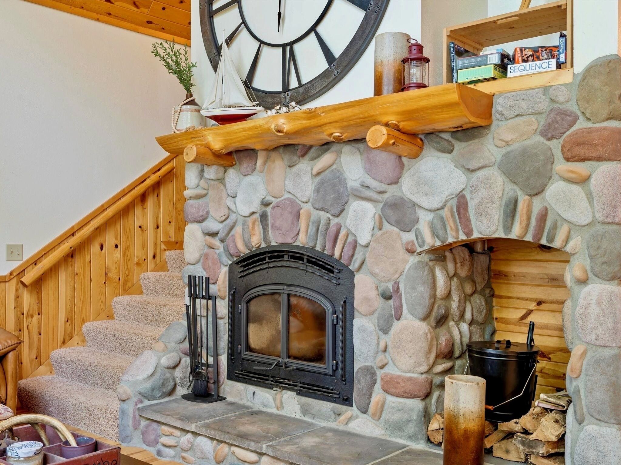 A stone fireplace with a clock above it, a wooden staircase, and a shelf with books and a lantern.