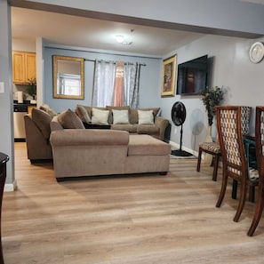 Un salon avec un canapé beige, une table à manger avec des chaises, un téléviseur et un ventilateur.
