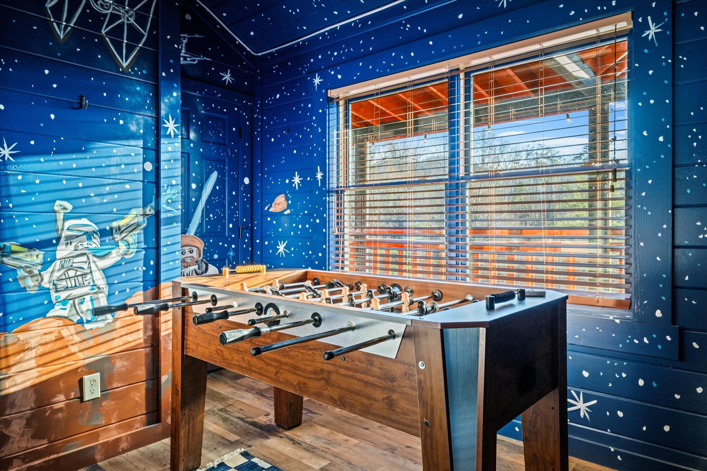 Una mesa de fútbolín en una habitación con paredes decoradas con motivos de estrellas y una ventana con persianas.