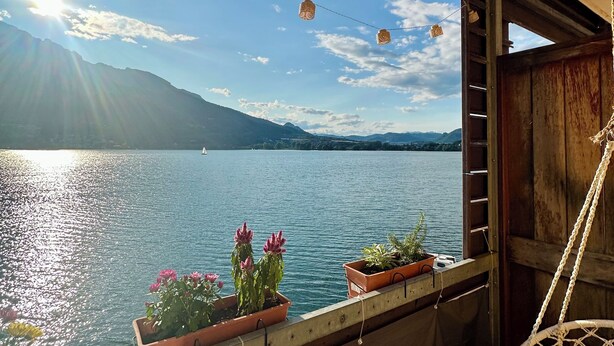 Un balcón con plantas en macetas, una silla colgante y vistas a un lago con montañas al fondo.
