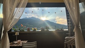 Un balcón con vistas a montañas y al sol poniéndose. Hay una silla, una mesa, una lámpara y una planta en maceta.