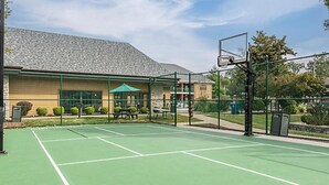 An outdoor tennis court with a basketball hoop, picnic tables, and a building with a green roof in the background. - BLOCK Falls Village 1br (Angels Camp)