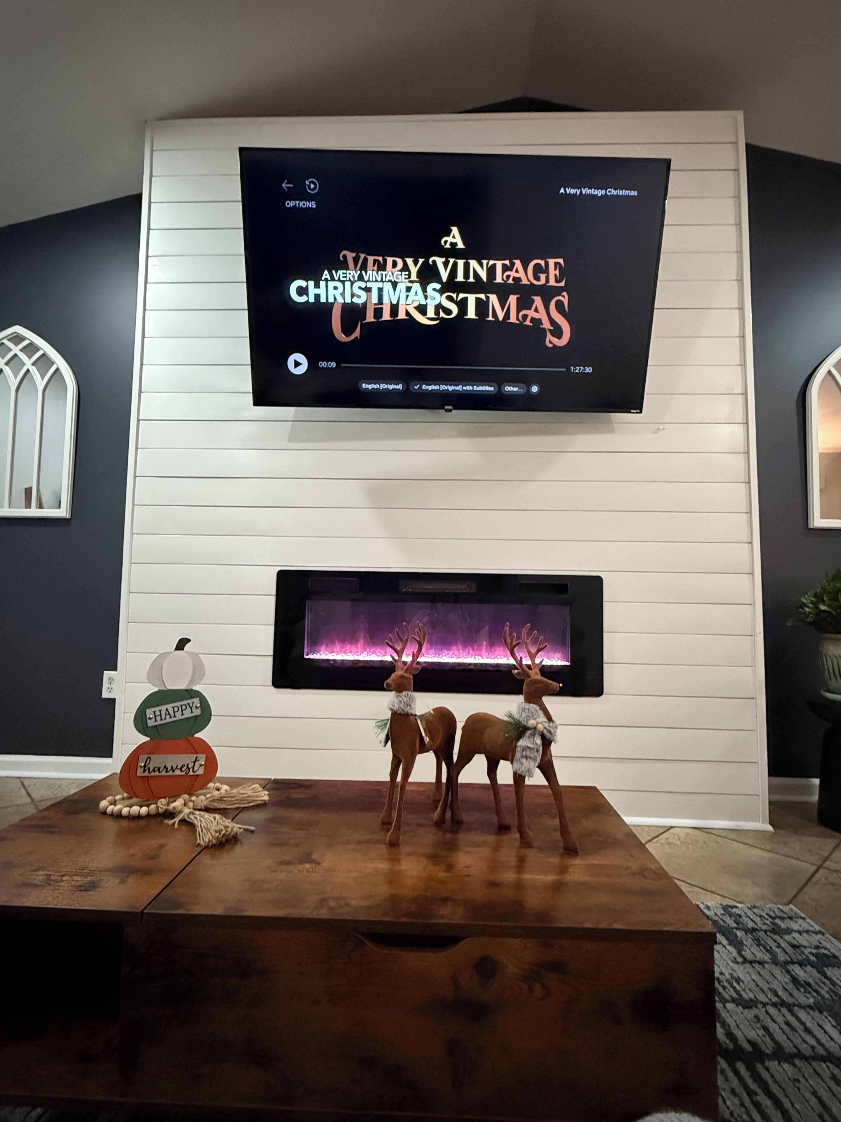A living room with a flat-screen TV mounted on a white wall, two deer figurines on a wooden coffee table, and a fireplace below the TV.