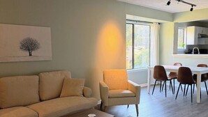 En stue med sofa, lænestol, sofabord og et tæppe med mønster. Der er et spiseområde med stole og et bord. Et vindue giver naturligt lys, og et køkken ses i baggrunden.