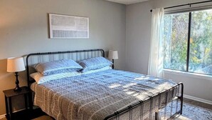 Et soveværelse med sengegavl i metal, et mønstret sengetæppe, et tæppe, et natbord med lampe og et vindue med gardiner.
