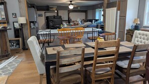 A dining area with a wooden table and chairs, a kitchen in the background, and a window with curtains. - large ski ranch hot tub close to killington pets ok (Mendon)